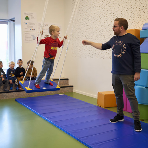 En dreng står på en gynge, som hænger i legerummet i børnehaven. En mandlig pædagogisk assistent står ved siden af og holder øjenkontakt med drengen.