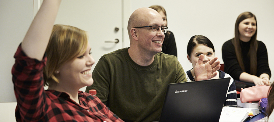 Billedet er taget i et undervisningslokale. I forgrunden sidder en kvindelig elev iført en ternet skjorte i rød og sort. Hun rækker højre hånd op og smiler bredt. bag hende sidder en mandlig elev iført en grøn bluse. Han har en opklappet pc stående foran sig, og han er i dialog med en person uden for billedet.
