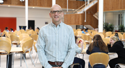 Claus Lundholm står på skoletorvet med elever og borde i baggrunden iført en blå skjorte.